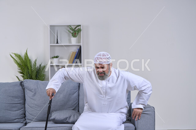 Aging and the need to use assistive devices, facial expressions indicating distress and pain, the need to go to the doctor and get treatment, portrait of an elderly Arab Gulf Emirati man wearing a kandura and a hat trying to use a medical crutch with expressions of pain, white background