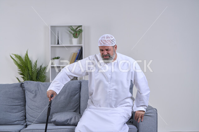 أهمية وضرورة الذهاب للطبيب والحصول على العلاج، التقدم في العمر والحاجه الى استخدام الوسائل المساعده، إيماءات الوجه تدل على الاضطراب والتألم، بورتريه لرجل كبير السن عربي خليجي إماراتي يرتدي الكندورة والقبعة يحاول القيام بمساعدة العكاز الطبي بتعابير الألم, خلفية بيضاء