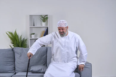 The importance and necessity of going to the doctor and getting treatment, aging and the need to use assistive devices, facial expressions indicating distress and pain, portrait of an elderly Arab Gulf Emirati man wearing a kandura and a hat trying to use a medical crutch with expressions of pain, white background