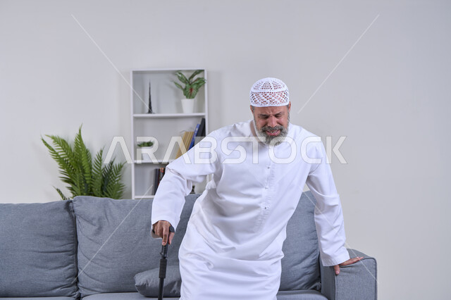 التقدم في العمر والحاجه الى استخدام الوسائل المساعده، إيماءات الوجه تدل على الاضطراب والتألم، ضرورة الذهاب للطبيب والحصول على العلاج، بورتريه لرجل كبير السن عربي خليجي إماراتي يرتدي الكندورة والقبعة يحاول القيام بمساعدة العكاز الطبي بتعابير الألم, خلفية بيضاء