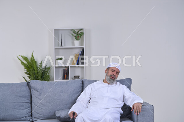 الاحساس بالتعب والوهن العام، ضرورة الذهاب للطبيب والحصول على العلاج، التقدم في العمر والحاجه الى المساعده، إيماءات الوجه تدل على الاضطراب والتألم، بورتريه لرجل كبير السن عربي خليجي إماراتي يرتدي الكندورة والقبعة يجلس على كنبه بتعابير الألم, خلفية بيضاء
