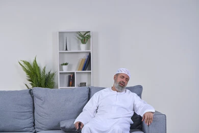 Feeling tired and general weakness, the need to go to the doctor and get treatment, aging and the need for help, facial expressions indicating distress and pain, portrait of an elderly Arab Gulf Emirati man wearing a kandura and a hat sitting on a sofa with an expression of pain, white background