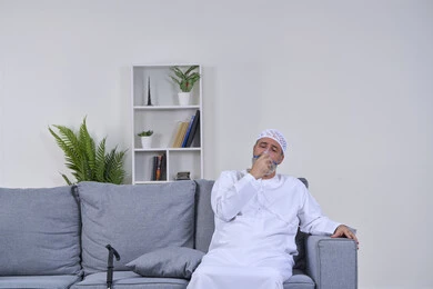 Maintaining a regular airway using a ventilator, portrait of an elderly Arab Gulf Emirati man wearing a kandura and hat sitting on a sofa and inhaling medication to treat sleep apnea through an oxygen mask, treatment of chronic asthma, white background