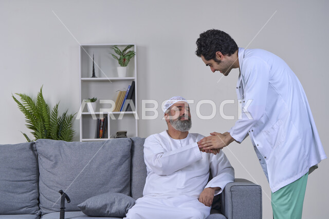 شركات التمرض المنزلي، الحاجة إلى العلاج الفيزيائي، آلام المفاصل والعظام والأعصاب، تقديم أفضل الخدمات الطبية المنزلية للمرضى كبار السن، بورتريه لممرض يرتدي المعطف يقوم بفحص رجل مسن عربي خليجي اماراتي يرتدي الكندورة ويجلس على الكنبه، خلفية بيضاء