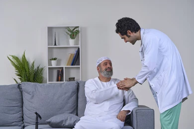 Home nursing companies, need for physical therapy, joint, bone and nerve pain, providing the best home medical services for elderly patients, portrait of a nurse wearing a coat examining an elderly Arab Gulf Emirati man wearing a kandura and sitting on a sofa, white background