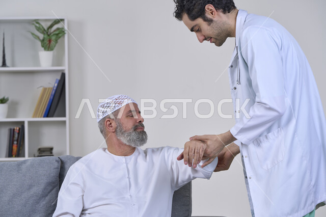 الحاجة إلى العلاج الفيزيائي، آلام المفاصل والعظام والأعصاب، شركات التمرض المنزلي، تقديم أفضل الخدمات الطبية المنزلية للمرضى كبار السن، بورتريه لممرض يرتدي المعطف يقوم بفحص رجل مسن عربي خليجي اماراتي يرتدي الكندورة ويجلس على الكنبه، خلفية بيضاء
