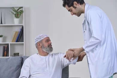 الحاجة إلى العلاج الفيزيائي، آلام المفاصل والعظام والأعصاب، شركات التمرض المنزلي، تقديم أفضل الخدمات الطبية المنزلية للمرضى كبار السن، بورتريه لممرض يرتدي المعطف يقوم بفحص رجل مسن عربي خليجي اماراتي يرتدي الكندورة ويجلس على الكنبه، خلفية بيضاء