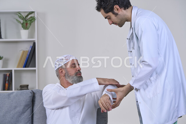 Joint, bone and nerve pain, facial expressions indicating distress and pain, home nursing companies, providing the best home medical services for elderly patients, portrait of a nurse wearing a coat examining an elderly Arab Gulf Emirati man wearing a kandura and sitting on a sofa, white background