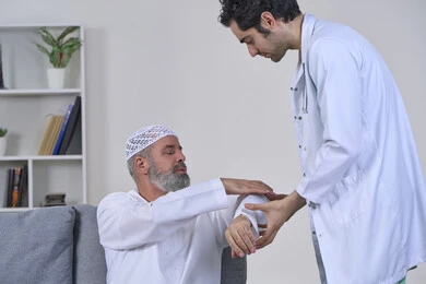 Joint, bone and nerve pain, facial expressions indicating distress and pain, home nursing companies, providing the best home medical services for elderly patients, portrait of a nurse wearing a coat examining an elderly Arab Gulf Emirati man wearing a kandura and sitting on a sofa, white background
