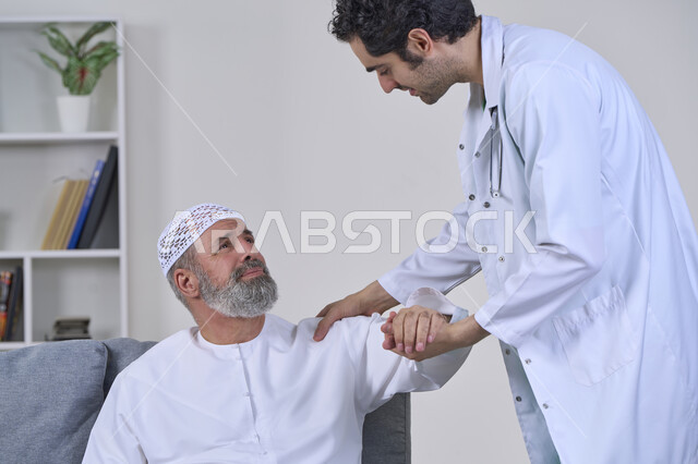 الحاجة إلى العلاج الفيزيائي، آلام المفاصل والعظام والأعصاب، شركات التمرض المنزلي، تقديم أفضل الخدمات الطبية المنزلية للمرضى كبار السن، بورتريه لممرض يرتدي المعطف يقوم بفحص رجل مسن عربي خليجي اماراتي يرتدي الكندورة ويجلس على الكنبه، خلفية بيضاء