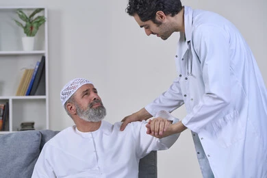 الحاجة إلى العلاج الفيزيائي، آلام المفاصل والعظام والأعصاب، شركات التمرض المنزلي، تقديم أفضل الخدمات الطبية المنزلية للمرضى كبار السن، بورتريه لممرض يرتدي المعطف يقوم بفحص رجل مسن عربي خليجي اماراتي يرتدي الكندورة ويجلس على الكنبه، خلفية بيضاء