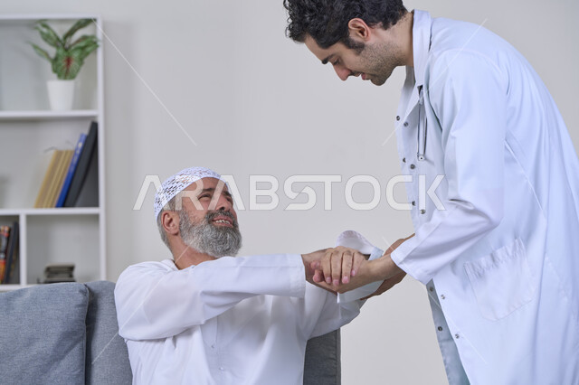 Joint, bone and nerve pain, facial expressions indicating distress and pain, home nursing companies, providing the best home medical services for elderly patients, portrait of a nurse wearing a coat examining an elderly Arab Gulf Emirati man wearing a kandura and sitting on a sofa, white background