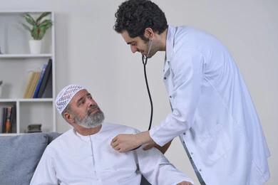 ايماءات الألم الشديد، استخدام السماعة الطبية لفحص القلب، شركات التمرض المنزلي، تقديم أفضل الخدمات الطبية المنزلية للمرضى كبار السن، بورتريه لممرض يرتدي المعطف يقوم بفحص رجل مسن عربي خليجي اماراتي يرتدي الكندورة ويجلس على الكنبه، خلفية بيضاء