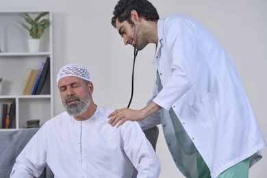 آلام الظهر والرقبة والعمود الفقري، شركات التمرض المنزلي، الحاجة إلى العلاج الفيزيائي، تقديم أفضل الخدمات الطبية المنزلية للمرضى كبار السن، بورتريه لممرض يرتدي المعطف يقوم بفحص رجل مسن عربي خليجي اماراتي يرتدي الكندورة ويجلس على الكنبه، خلفية بيضاء