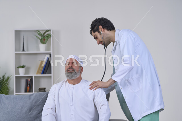 استخدام السماعة الطبية، شركات التمرض المنزلي، آلام الظهر والعمود الفقري، الحاجة إلى العلاج الفيزيائي، تقديم أفضل الخدمات الطبية المنزلية للمرضى كبار السن، بورتريه لممرض يرتدي المعطف يقوم بفحص رجل مسن عربي خليجي اماراتي يرتدي الكندورة ويجلس على الكنبه، خلفية بيضاء