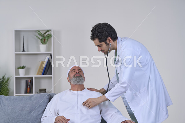 Severe pain gestures, using a stethoscope to examine the heart, home nursing companies, providing the best home medical services for elderly patients, portrait of a nurse wearing a coat examining an elderly Arab Gulf Emirati man wearing a kandura and sitting on a sofa, white background