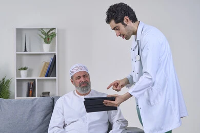 A nurse wearing a uniform and coat describing the patient's condition using a tablet device, providing the best home medical services for elderly patients, following up on the patient's electronic medical record, a portrait of an elderly Arab Gulf Emirati patient wearing a kandura and sitting on a sofa, white background