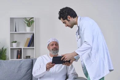 تقديم أفضل الخدمات الطبية المنزلية لمرضى كبار السن، متابعة السجل الطبي الالكتروني الخاص بالمريض, بورتريه لممرض يرتدي اليونيفورم والمعطف يقوم بوصف الحالة المرضية باستخدام جهاز التابليت، مريض مسن عربي خليجي اماراتي يرتدي الكندورة ويجلس على الكنبه، خلفية بيضاء
