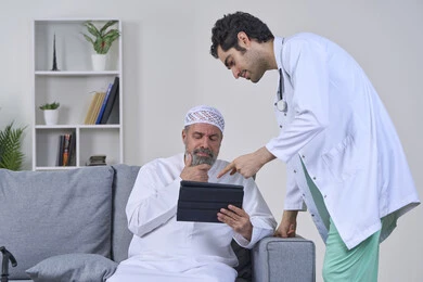 Providing the best home medical services for elderly patients, monitoring the patient's electronic medical record, portrait of a nurse wearing a uniform and coat describing the patient's condition using a tablet, an elderly Arab Gulf Emirati patient wearing a kandura and sitting on a sofa, white background