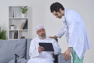 متابعة السجل الطبي الالكتروني الخاص بالمريض, ممرض يرتدي اليونيفورم والمعطف يقوم بوصف الحالة المرضية باستخدام جهاز التابليت، تقديم أفضل الخدمات الطبية المنزلية لمرضى كبار السن، بورتريه لرجل مسن عربي خليجي اماراتي يرتدي الكندورة ويجلس على الكنبه، خلفية بيضاء