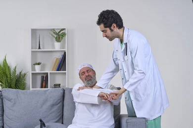 Joint, bone and nerve pain, facial expressions indicating distress and pain, home nursing companies, providing the best home medical services for elderly patients, portrait of a nurse wearing a coat examining an elderly Arab Gulf Emirati man wearing a kandura and sitting on a sofa, white background