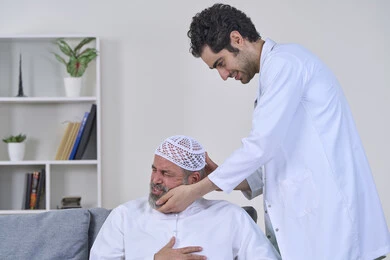 Home nursing companies, back, neck and spine pain, need for physical therapy, providing the best home medical services for elderly patients, portrait of a nurse wearing a coat examining an elderly Arab Gulf Emirati man wearing a kandura and sitting on a sofa, white background