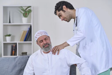 Back, neck and spine pain, home nursing companies, need for physical therapy, providing the best home medical services for elderly patients, portrait of a nurse wearing a coat examining an elderly Arab Gulf Emirati man wearing a kandura and sitting on a sofa, white background