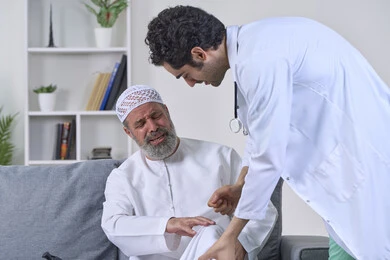 شركات التمرض المنزلي، الحاجة إلى العلاج الفيزيائي، آلام المفاصل والعظام والأعصاب، تقديم أفضل الخدمات الطبية المنزلية للمرضى كبار السن، بورتريه لممرض يرتدي المعطف يقوم بفحص رجل مسن عربي خليجي اماراتي يرتدي الكندورة ويجلس على الكنبه، خلفية بيضاء