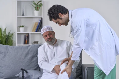 الحاجة إلى العلاج الفيزيائي، آلام المفاصل والعظام والأعصاب، شركات التمرض المنزلي، تقديم أفضل الخدمات الطبية المنزلية للمرضى كبار السن، بورتريه لممرض يرتدي المعطف يقوم بفحص رجل مسن عربي خليجي اماراتي يرتدي الكندورة ويجلس على الكنبه، خلفية بيضاء