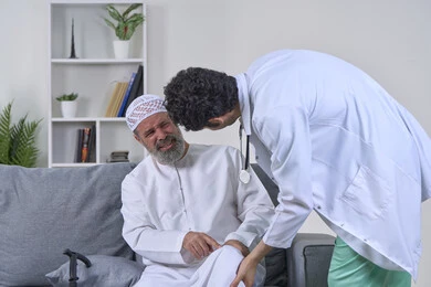 شركات التمرض المنزلي، الحاجة إلى العلاج الفيزيائي، آلام المفاصل والعظام والأعصاب، تقديم أفضل الخدمات الطبية المنزلية للمرضى كبار السن، بورتريه لممرض يرتدي المعطف يقوم بفحص رجل مسن عربي خليجي اماراتي يرتدي الكندورة ويجلس على الكنبه، خلفية بيضاء