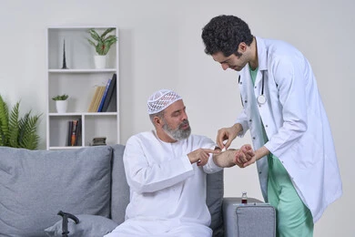 إعطاء حقنة مسكنة للألم، العمل في شركات التمريض المنزلي، تقديم أفضل الخدمات الطبية المنزلية للمرضى كبار السن، بورتريه لممرض يرتدي اللباس الطبي يقوم يتعقيم مكان الابرة لرجل كبير بالسن عربي خليجي اماراتي يرتدي الكندورة ويجلس على كنبه، خلفية بيضاء