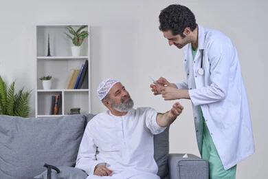 Preparing for a painkiller injection, the concept of working in home nursing companies, providing the best home medical services for elderly patients, a portrait of a nurse wearing medical clothing giving an injection to an elderly Arab Gulf Emirati man wearing a kandura and sitting on a sofa, white background
