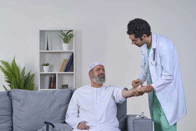 أخذ الحقنة المسكنة للوجع، تعابير الألم الشديد، تقديم أفضل الخدمات الطبية المنزلية للمرضى كبار السن، العمل في القطاع الصحي، بورتريه لممرض اسعافات اولية يرتدي اللباس الطبي يقوم بإعطاء ابرة لرجل كبير بالسن عربي خليجي اماراتي يرتدي الكندورة ويجلس على كنبه، خلفية بيضاء