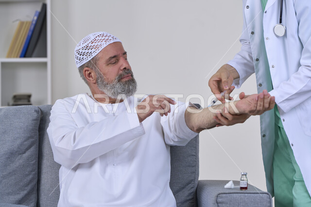 The concept of working in home nursing companies, taking a painkiller injection, providing the best home medical services for elderly patients, close-up portrait of a first aid nurse wearing medical clothing giving an injection to an elderly Arab Gulf Emirati man wearing a kandura and sitting on a sofa, white background
