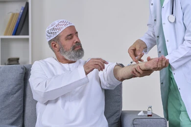 The concept of working in home nursing companies, taking a painkiller injection, providing the best home medical services for elderly patients, close-up portrait of a first aid nurse wearing medical clothing giving an injection to an elderly Arab Gulf Emirati man wearing a kandura and sitting on a sofa, white background