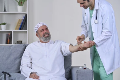 تعابير الألم الشديد، شركات التمرض المنزلي، تقديم أفضل الخدمات الطبية المنزلية للمرضى كبار السن، العمل في القطاع الصحي، بورتريه لممرض اسعافات اولية يرتدي اليونيفورم الطبي يقوم بإعطاء ابرة لرجل كبير بالسن عربي خليجي اماراتي يرتدي الكندورة ويجلس على كنبه، خلفية بيضاء