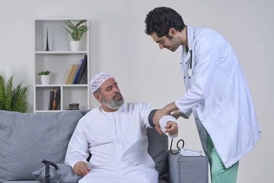 تقديم الرعاية الصحية المنزلية، الفحص الطبي والتشخيص السريع، متابعة دورية لفحص الضغط، العمل في القطاع الصحي، بورتريه لممرض يرتدي المعطف ويضع السماعة الطبية يقوم بقياس ضغط الدم لمريض عربي خليجي إماراتي يرتدي الكندورة والقبعة يجلس على الكنبه، خلفية بيضاء