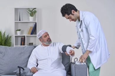 الفحص الطبي والتشخيص السريع، تقديم الرعاية الصحية المنزلية، متابعة دورية لفحص الضغط، العمل في القطاع الصحي، بورتريه لممرض يرتدي المعطف ويضع السماعة الطبية يقوم بقياس ضغط الدم لمريض عربي خليجي إماراتي يرتدي الكندورة والقبعة يجلس على الكنبه، خلفية بيضاء