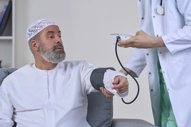Periodic follow-up for blood pressure check, providing home health care, medical examination and rapid diagnosis, working in the health sector, close-up portrait of a nurse wearing a medical coat measuring the blood pressure of an Arab Gulf Emirati patient wearing a kandura and a hat sitting on a sofa, white background