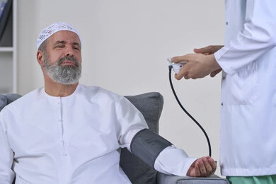 Feeling pain and soreness, regular follow-up to check blood pressure, providing home health care, medical examination and rapid diagnosis, working in the health sector, close-up portrait of a nurse wearing a coat measuring the blood pressure of an Arab Gulf Emirati patient wearing a kandura and hat sitting on a sofa, white background