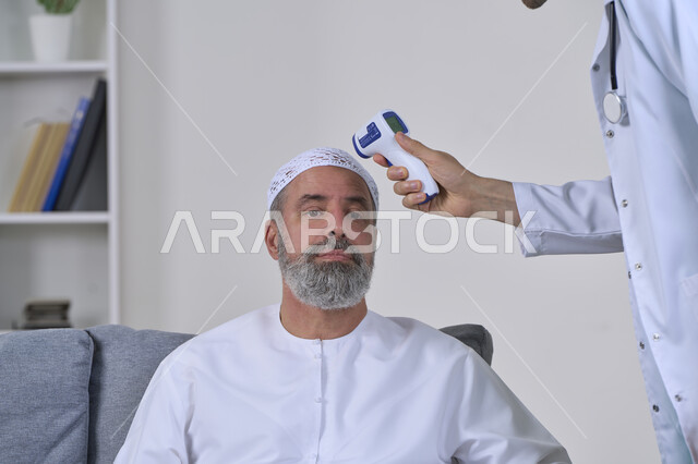طبيب يحمل مقياس الحرارة الرقمي بالأشعة تحت الحمراء، إتباع إجراءات الوقاية و السلامة من فايروس كورونا، الوعي الصحي وتجنب إنتشار العدوى، بورتريه مقرب لممرض يقوم بفحص مريض مسن عربي خليجي اماراتي يرتدي الكندورة والقبعة، العمل في القطاع الطبي، خلفية بيضاء