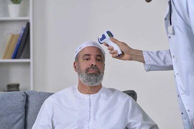 A doctor holding a digital infrared thermometer, following preventive and safety measures against the Corona virus, health awareness and avoiding the spread of infection, close-up portrait of a nurse examining an elderly Arab Gulf Emirati patient wearing a kandura and a hat, working in the medical sector, white background