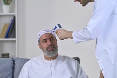 إتباع إجراءات الوقاية و السلامة من فايروس كورونا، طبيب يحمل مقياس الحرارة الرقمي بالأشعة تحت الحمراء، الوعي الصحي وتجنب إنتشار العدوى، بورتريه مقرب لممرض يقوم بفحص مريض مسن عربي خليجي اماراتي يرتدي الكندورة والقبعة، العمل في القطاع الطبي، خلفية بيضاء