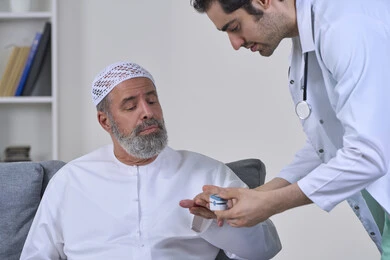 Periodic follow-up of home checkups, doctor in coat holding digital infrared pulse oximeter, close-up portrait of nurse checking blood oxygen level of elderly Arab Gulf Emirati patient wearing kandura and hat, working in medical sector, white background
