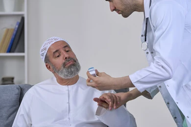 Pulse oximeter, home checkup follow-up, doctor in coat holding digital infrared pulse oximeter, close-up portrait of nurse examining elderly Arab Gulf Emirati patient wearing kandura and hat, working in medical sector, white background