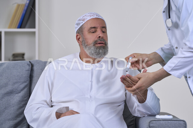 جهاز قياس سكر الدم لمرضى السكري، العمل في القطاع الطبي وشركات التمريض المنزلي، الرعاية الصحية والفحص الطبي السريع، بورتريه مقرب لممرض يرتدي المعطف الطبي يقوم بفحص نسبة السكر في الدم لمريض مسن عربي خليجي اماراتي يرتدي الكندورة يجلس على الكنبه، خلفية بيضاء