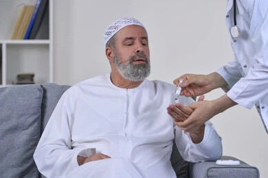 Blood glucose meter for diabetics, working in the medical sector and home nursing companies, healthcare and rapid medical examination, close-up portrait of a nurse wearing a medical coat checking the blood sugar level of an elderly Arab Gulf Emirati patient wearing a kandura sitting on a sofa, white background