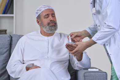 العمل في القطاع الطبي وشركات التمريض المنزلي، الرعاية الصحية والفحص الطبي السريع، بورتريه مقرب لممرض يرتدي المعطف الطبي يقوم بفحص نسبة السكر في الدم لمريض مسن عربي خليجي اماراتي يرتدي الكندورة يجلس على الكنبه، جهاز قياس سكر الدم لمرضى السكري، خلفية بيضاء