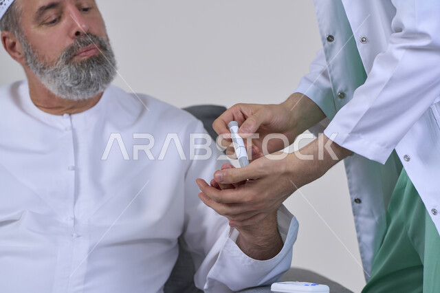جهاز قياس سكر الدم لمرضى السكري، العمل في القطاع الطبي وشركات التمريض المنزلي، الرعاية الصحية والفحص الطبي السريع، بورتريه مقرب لممرض يرتدي المعطف الطبي يقوم بفحص نسبة السكر في الدم لمريض مسن عربي خليجي اماراتي يرتدي الكندورة يجلس على الكنبه، خلفية بيضاء