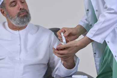 جهاز قياس سكر الدم لمرضى السكري، العمل في القطاع الطبي وشركات التمريض المنزلي، الرعاية الصحية والفحص الطبي السريع، بورتريه مقرب لممرض يرتدي المعطف الطبي يقوم بفحص نسبة السكر في الدم لمريض مسن عربي خليجي اماراتي يرتدي الكندورة يجلس على الكنبه، خلفية بيضاء